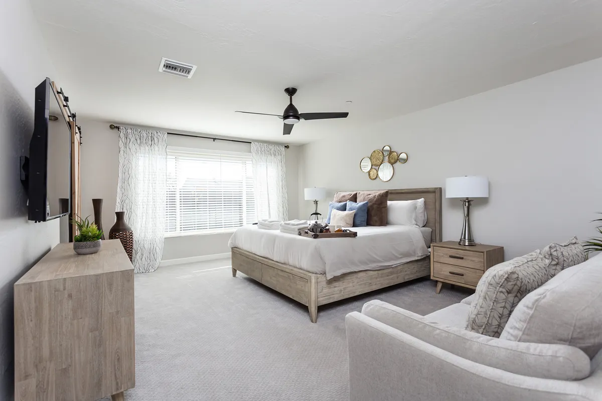 A spacious bedroom featuring a plush bed adorned with decorative pillows, a seating area with an armchair, a wall-mounted television, tasteful wall art, and bedside lamps. The overall aesthetic is tranquil and cozy, inviting relaxation and restful sleep.
