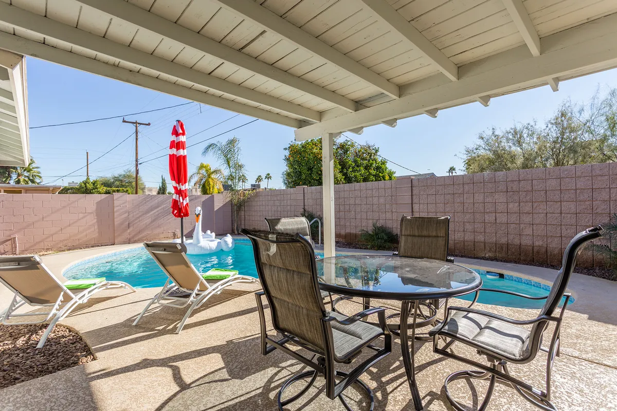 A tranquil poolside view with a swimming pool, red and white poolside umbrella, a dining table with chairs, and multiple lounge chairs arranged for sunbathing. The lighting suggests a relaxing afternoon or early evening mood, perfect for sunset cocktails or social gatherings.