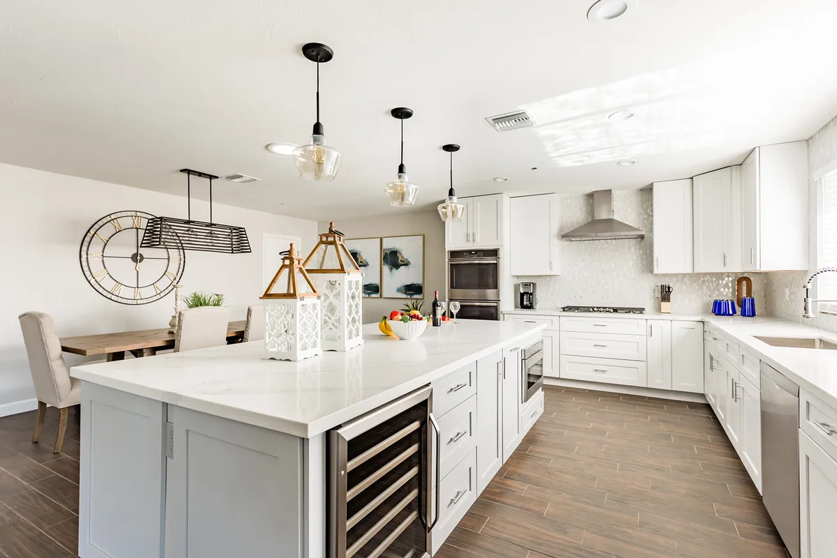 A modern kitchen is highlighted by a large island, stylish hanging light fixtures, a colorful fruit centerpiece, and a view of the adjacent dining area adorned with artwork. The open design encourages collaborative cooking and seamless socializing, while the dining area signals space for shared meals and memories.