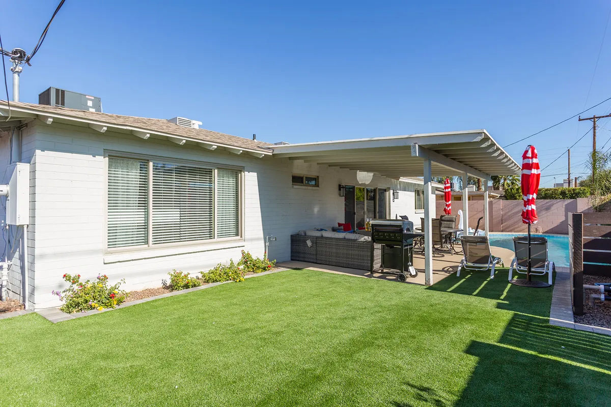 A cheerful backyard scene featuring a swimming pool, grill station, grassy lawn, outdoor seating area, and vibrant red umbrellas. The space is clearly designed for social gatherings and relaxation, with ample room for family or group activities.
