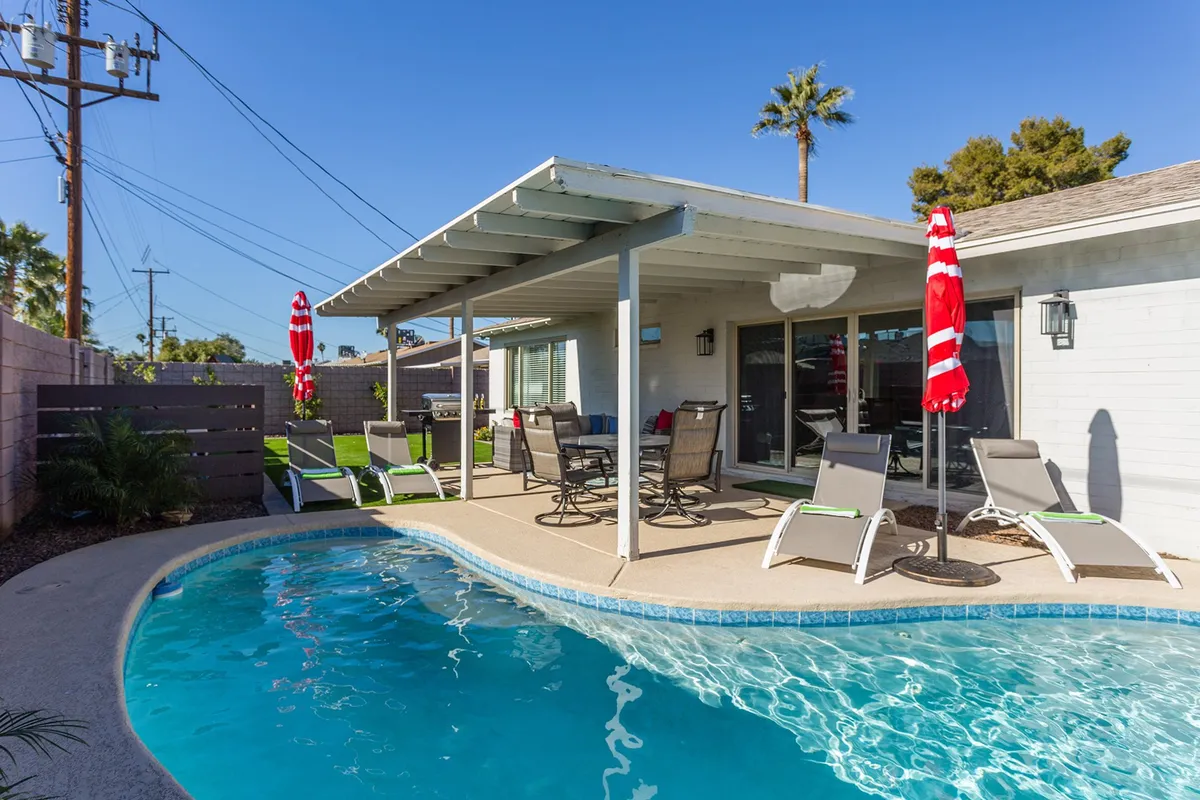 A sunny backyard scene featuring a sparkling swimming pool surrounded by a spacious patio. There are multiple lounge chairs for sunbathing, a shaded covered patio with an outdoor dining set, and greenery along the edges, creating a private resort-like atmosphere. The space looks inviting for both relaxation and social gatherings, with the pool as the central feature.