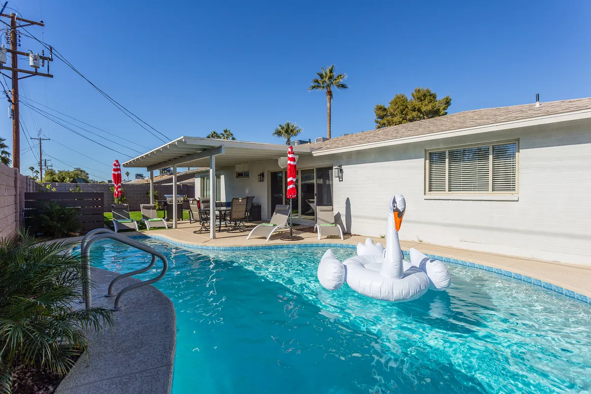 The image showcases a sparkling swimming pool with an eye-catching inflatable swan, surrounded by a patio area perfect for lounging, swaying palm trees, and inviting spaces set against the warm Scottsdale sky. The setup hints at playful pool days and vibrant gatherings, amplified by the sunset glow and the promise of outdoor leisure.