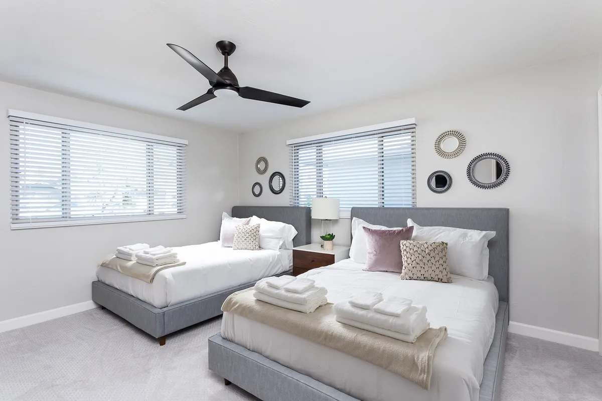 A guest bedroom showcasing two beds with stylish linens, decorative mirrors above the headboards, a window with soft treatments, and a nightstand with a lamp. The room exudes a calm and cozy mood, perfect for children or friends.