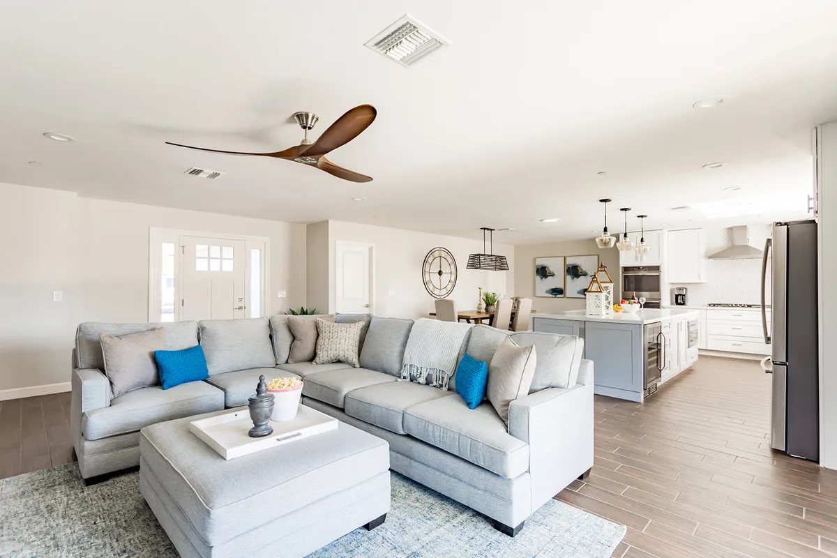 A comfortable interior scene displays a large sectional sofa with a coffee table, a modern ceiling fan overhead, and the kitchen area with an island visible in the background. The open flow between living and kitchen spaces highlights the home's social, entertainer-friendly design, perfect for gatherings and celebrations.